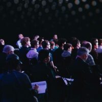 A diverse group of people sit closely together in rows of chairs, attentively facing forward in a dimly lit auditorium with a dark background and soft overhead lights.