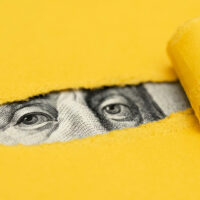A close-up of eyes from a U.S. dollar bill peeking through a tear in bright yellow paper, with the paper rolled back to reveal the eyes.