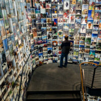 A person stands on a spiral staircase, looking at walls covered with hundreds of colorful photographs displayed in a brightly lit, modern gallery or exhibition space.