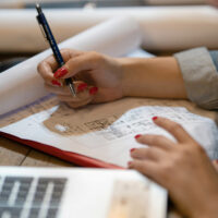 A person with red nail polish writes notes on architectural plans at a desk, with a laptop and rolled-up papers nearby.