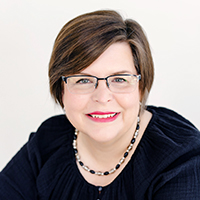 Woman with short brown hair wearing glasses, a black top, a beaded necklace, and red lipstick, smiling at the camera against a light background.