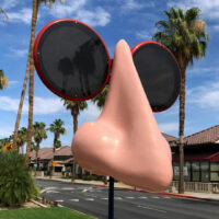 A large sculpture of a nose with oversized sunglasses stands on a street lined with palm trees and buildings under a blue sky.