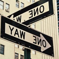 Two One Way street signs point in opposite directions on a city street, with tall buildings in the background.
