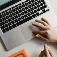 Person using a laptop on a white desk, with a drink, an orange book, and a small green potted plant nearby. Only hands are visible; one hand is on the laptops touchpad.