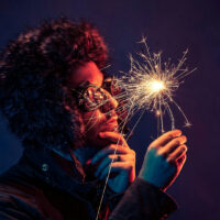 A person wearing a furry winter hat and glasses holds a lit sparkler, looking at it thoughtfully against a dark, colorful background.