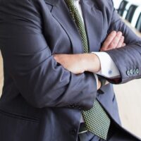 A person in a business suit stands with arms crossed at the head of a long, empty conference table in a modern office setting.